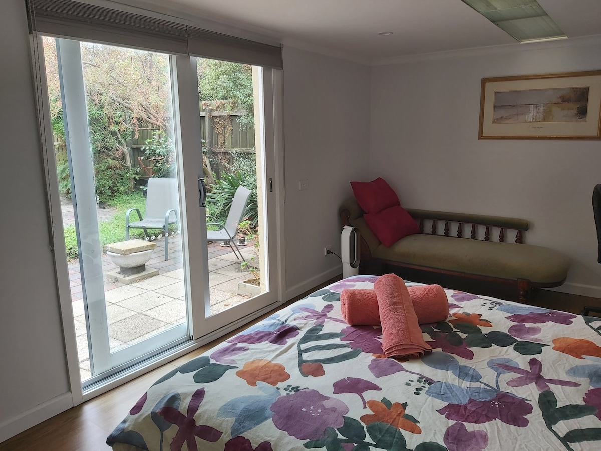 A spacious bedroom features a floral patterned quilt on a large bed. A daybed sits in the corner, and glass sliding doors open to a serene garden with outdoor seating and a stone fire pit.