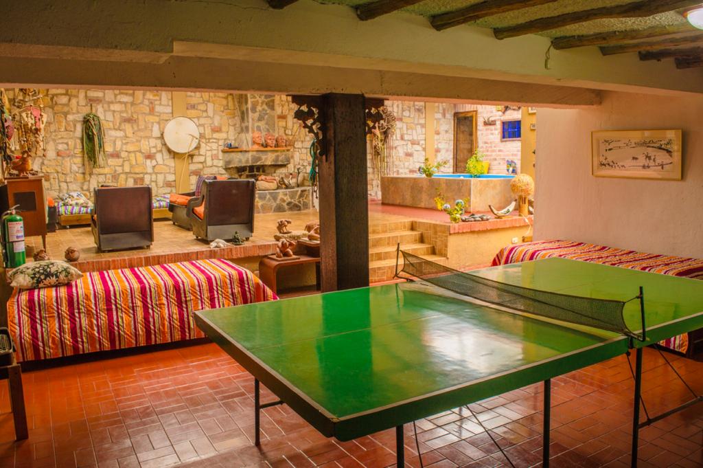 A spacious common area is highlighted by a green ping pong table, positioned between two striped sofa beds. A stone wall serves as a backdrop, accented by decorative sculptures and indoor plants. The area provides a glimpse into the cozy seating space with chairs in the background.