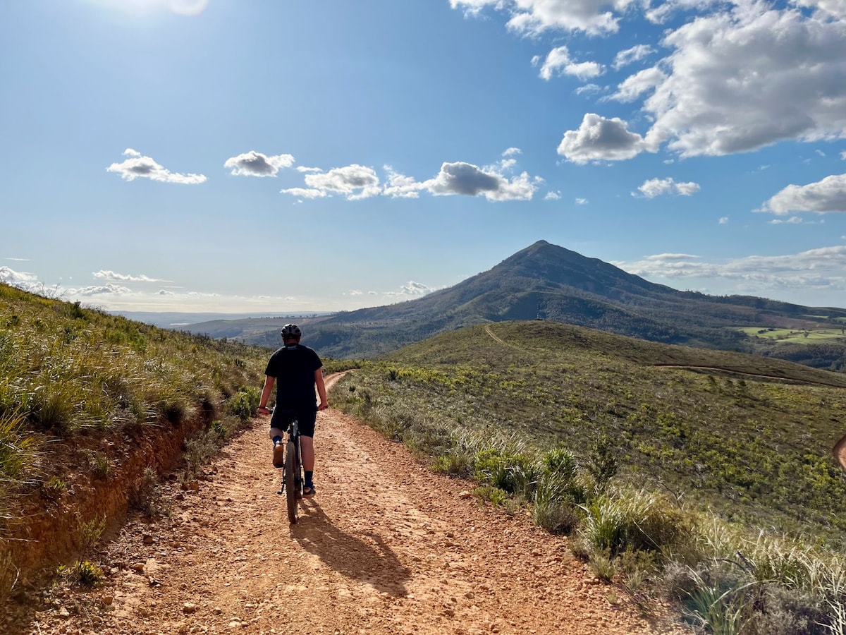 A cyclist is seen riding along a dirt trail that winds through a lush, hilly landscape. Mountains rise majestically in the distance, under a bright sky filled with scattered clouds. The path is bordered by verdant grass and offers a sense of serenity and natural beauty.