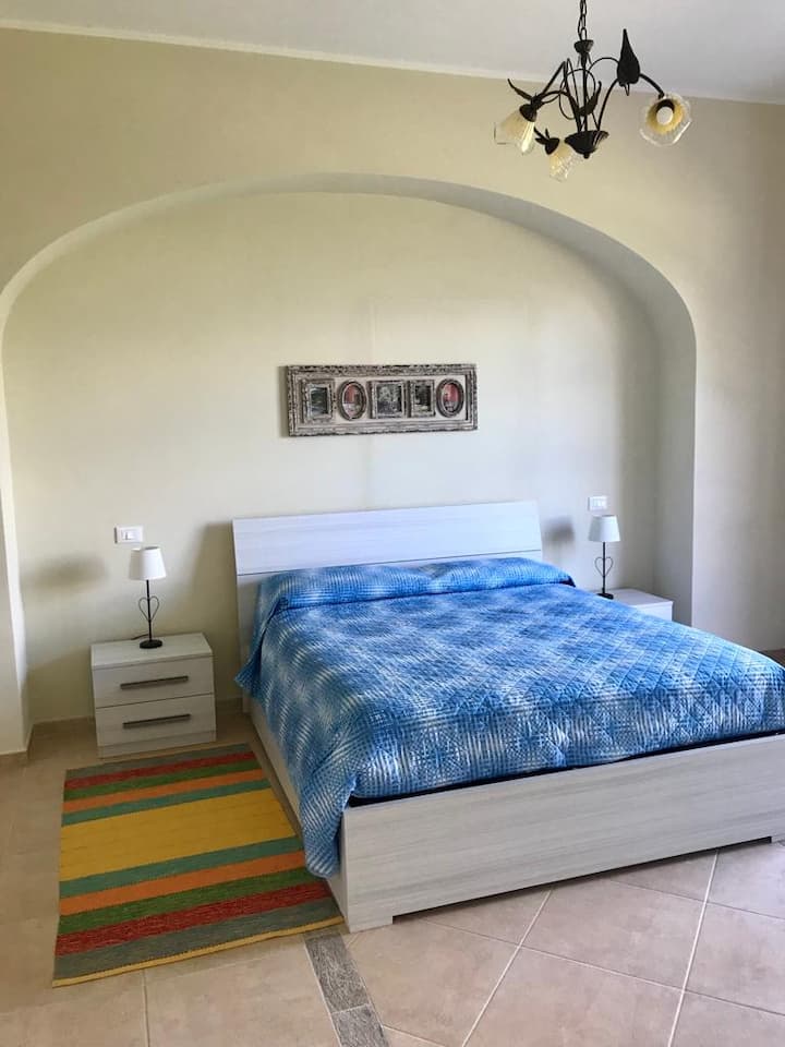 Dhoma e gjumit 1: Një dhomë gjumi me hapësirë të bollshme me një krevat dopio të madh në qendër. Muret janë të lyera me ngjyra relaksuese, si bezhë. Një qilim i butë mbulon dyshemenë, duke e bërë atë të rehatshme. 