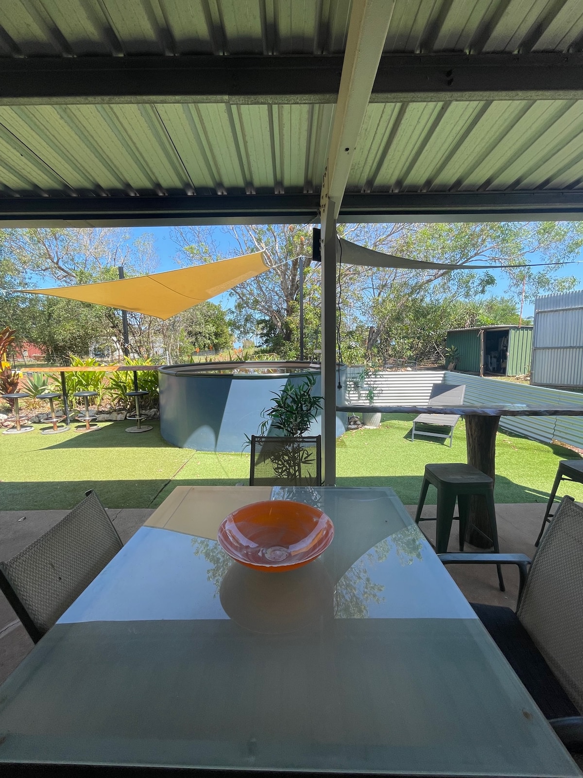 An outdoor dining area features a glass table with chairs, complemented by a decorative bowl at the center. Sun shades provide coverage, while a green lawn and various plants create a refreshing atmosphere in the background.