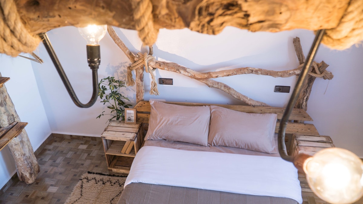 A cozy guest room features a spacious bed with a neutral-toned comforter and soft pillows. Natural wood elements, including a decorative wall structure, enhance the ambiance. Ambient lighting from hanging bulbs contributes to the soothing atmosphere, while a small plant adds a touch of greenery.
