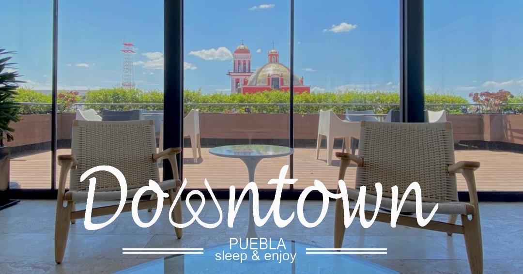 A spacious rooftop terrace is shown, featuring two comfortable chairs facing large glass doors. A panoramic view of Puebla's skyline is visible, with notable architectural elements in the distance. Sunlight brightens the scene, enhancing the overall openness of the space.