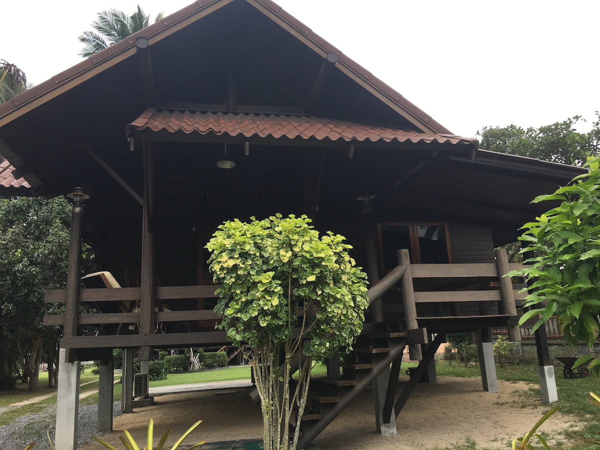 A two-story log home is presented with a sloped roof and a spacious balcony. A well-maintained garden with green foliage surrounds the entrance. Steps lead up to the porch, where outdoor seating is positioned. The house is set amid a serene landscaping.