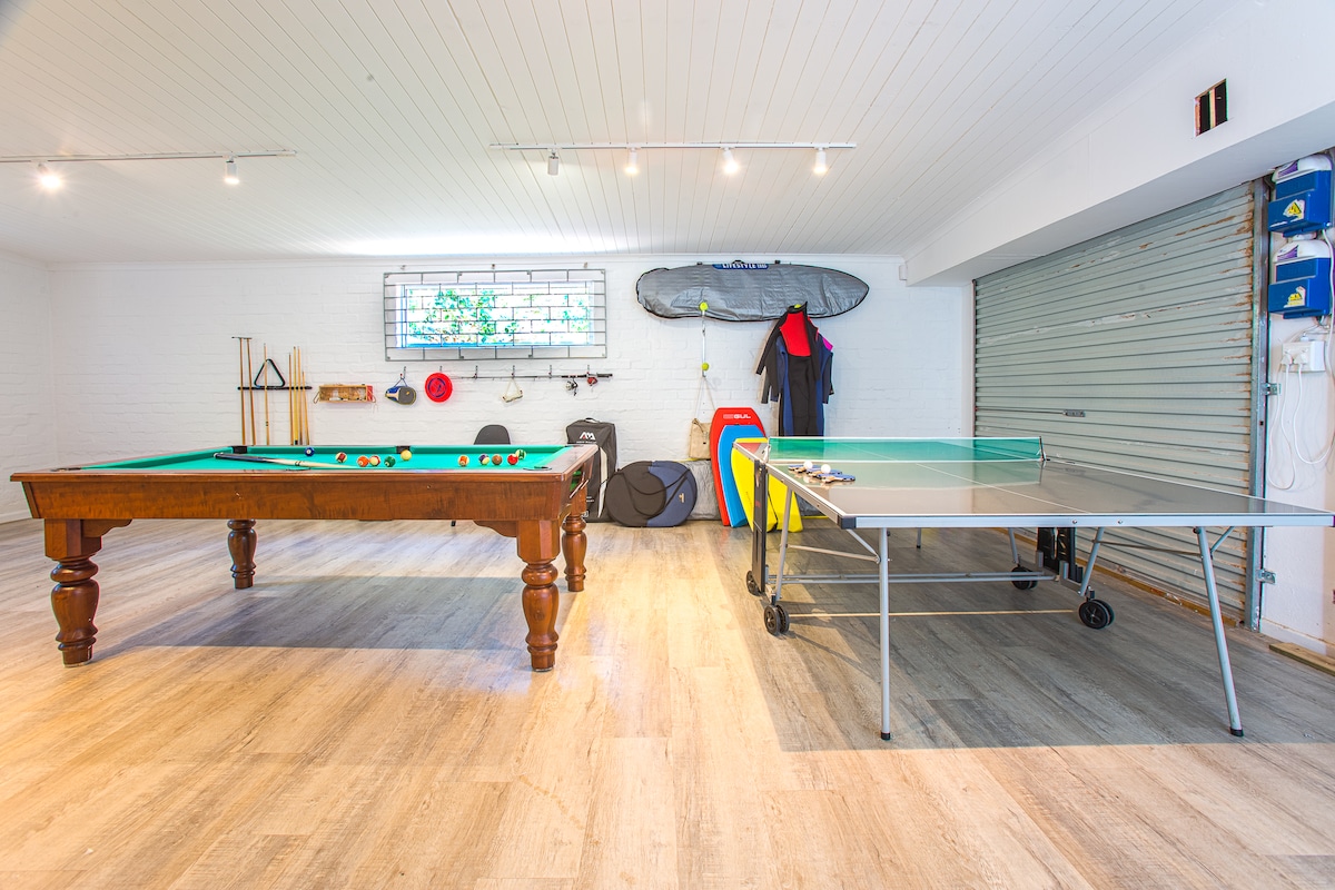 A games room is highlighted with a pool table and a ping-pong table, providing recreational options for guests. The walls are adorned with sports equipment, including surfboards. Ample lighting enhances the bright, open space, perfect for family entertainment.