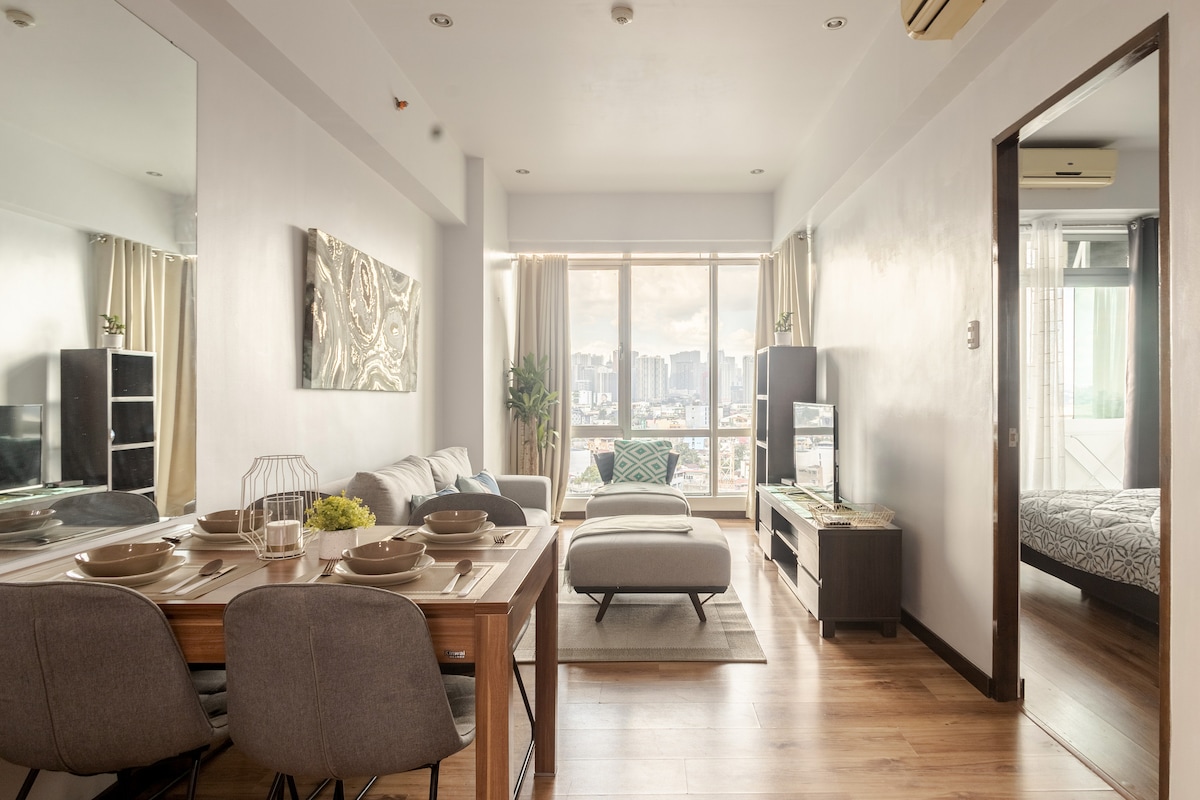 A spacious living area features large windows offering unobstructed city views. A dining table for four is set alongside a comfortable seating area, with neutral décor and natural light enhancing the ambiance. The adjoining bedroom is visible through an open door.