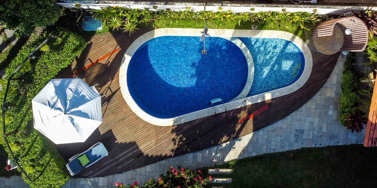 The image captures an aerial view of a two-level swimming pool, featuring vibrant blue water. Surrounding the pool, wooden decking offers space for lounging. A shaded umbrella and a small seating area are visible, along with lush greenery enhancing the outdoor atmosphere.