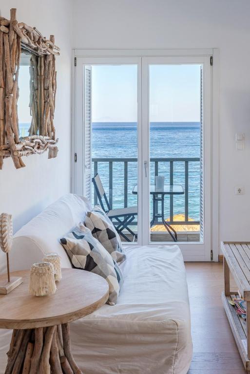 A light-filled living area features a white sofa adorned with patterned cushions. A rustic mirror made of driftwood reflects the serene view of the sea. Glass doors lead to a private balcony with a small table and chairs, offering an unobstructed ocean vista.
