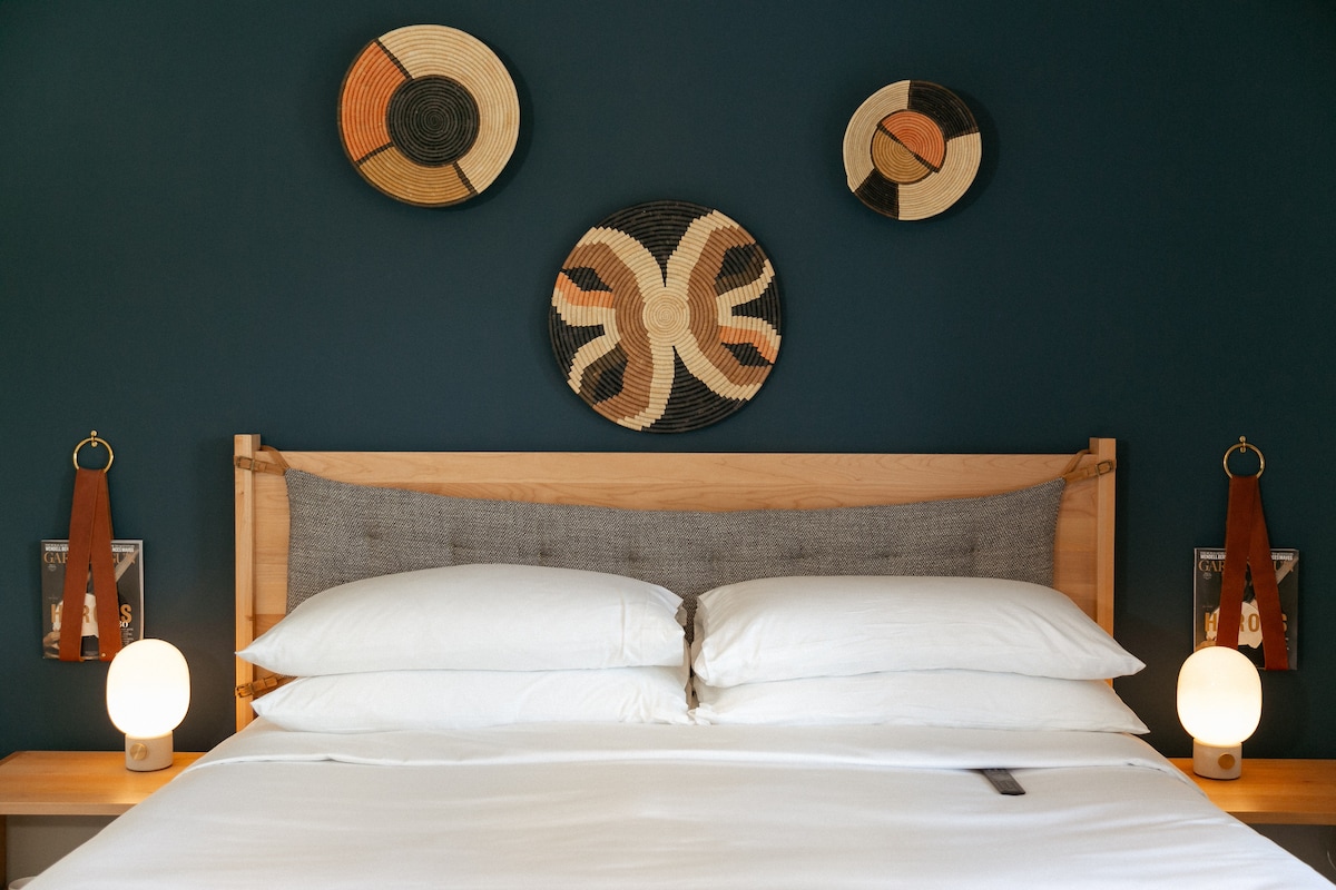 A cozy sleeping area showcases a spacious bed with crisp white linens. Elegant round wall art made from natural materials complements the dark blue backdrop, while two stylish bedside lamps provide soft illumination on either side.