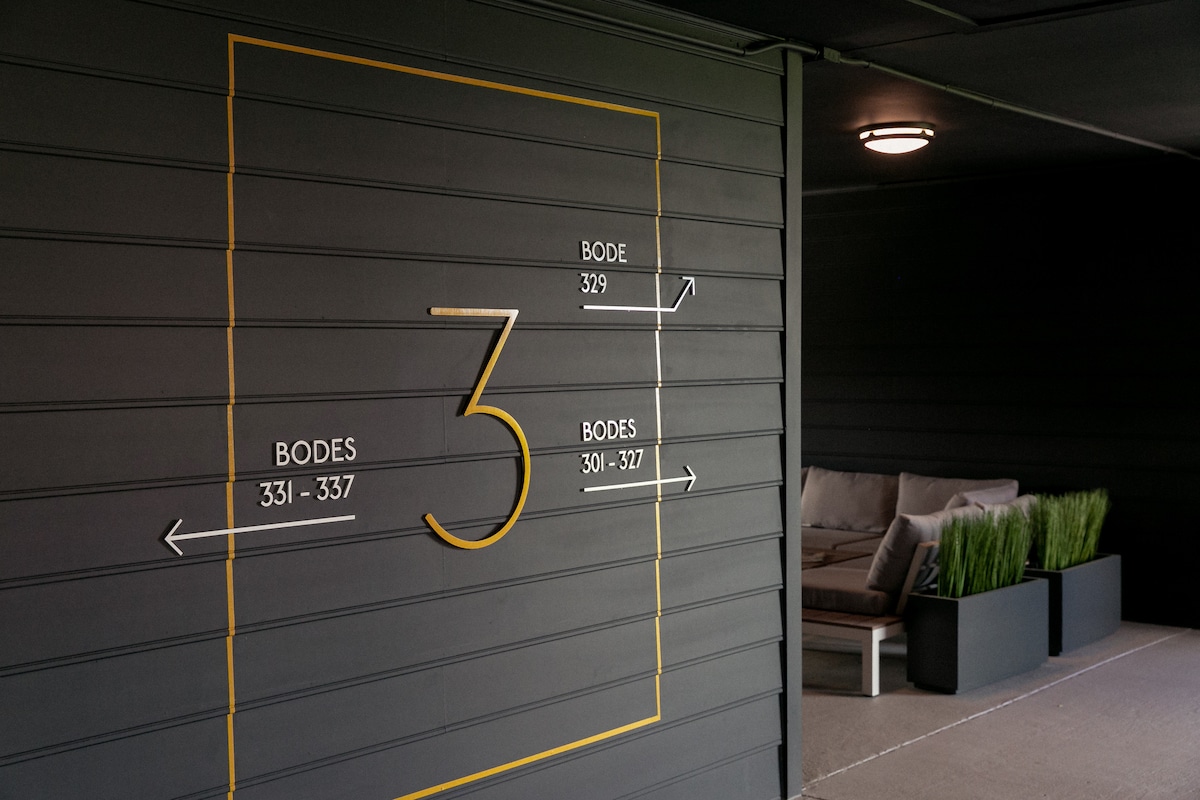 A signage area displays directional information and the number three, guiding guests to Bode Suites 301-337. The muted grey siding creates a subtle backdrop, while a seating area with soft cushions and planters adds a cozy touch to the entrance.