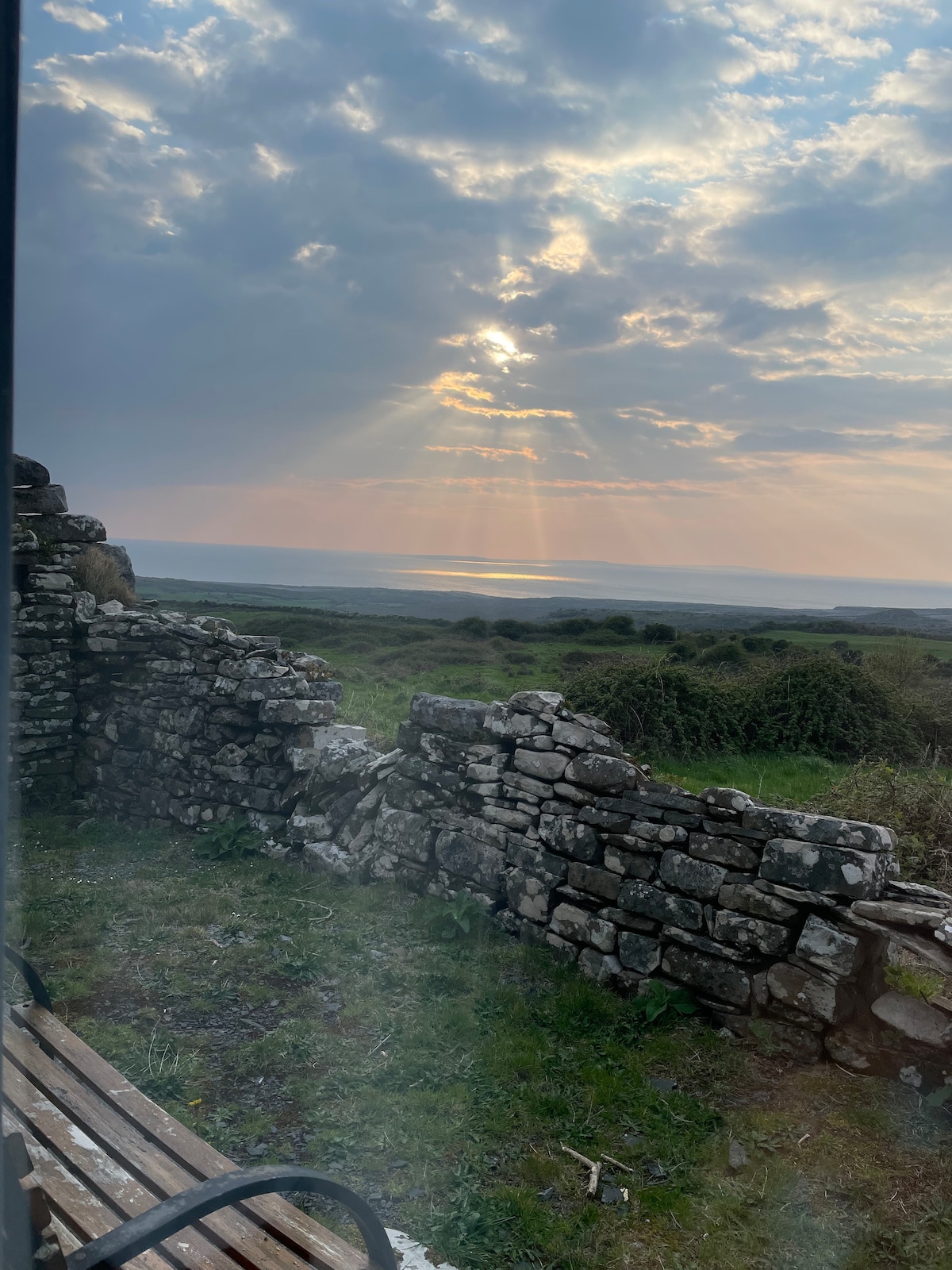 A partial view of a stone wall stands in front of a grassy area, overlooking the landscape. Sunlight beams through the cloud cover, illuminating the horizon, where the ocean is visible in the distance. The scene conveys a sense of tranquility and natural beauty.
