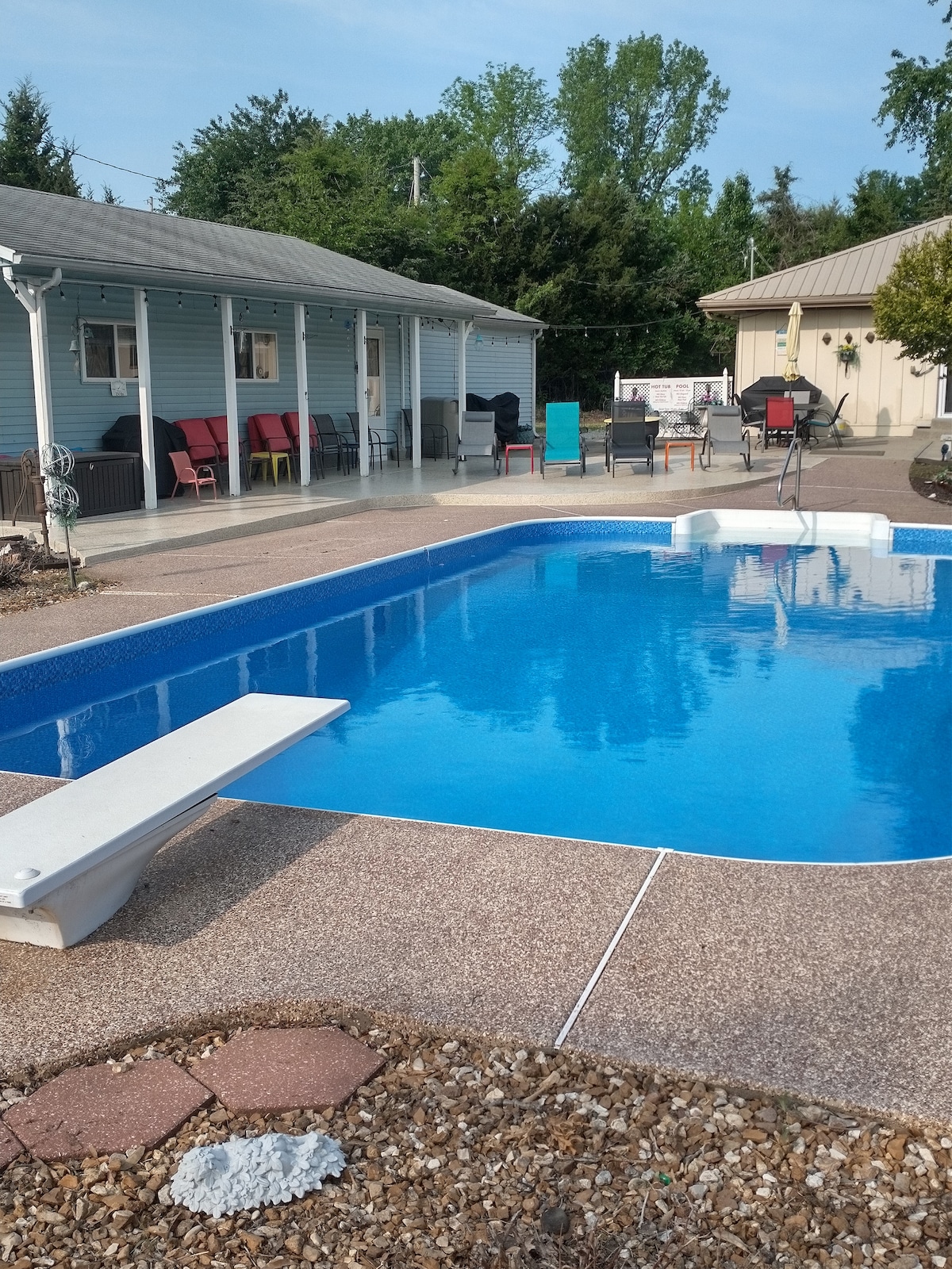 A clear blue saltwater pool is surrounded by a spacious patio featuring multiple outdoor seating areas. The guest house and additional structures can be seen in the background, with lounge chairs and tables providing a comfortable space for relaxation.