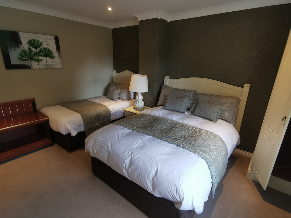 A spacious room is presented with a large double bed and a large single bed, both featuring soft bedding and decorative pillows. A bedside lamp stands next to the double bed, and a wooden bench is positioned against the wall. Subtle artwork adorns the wall.