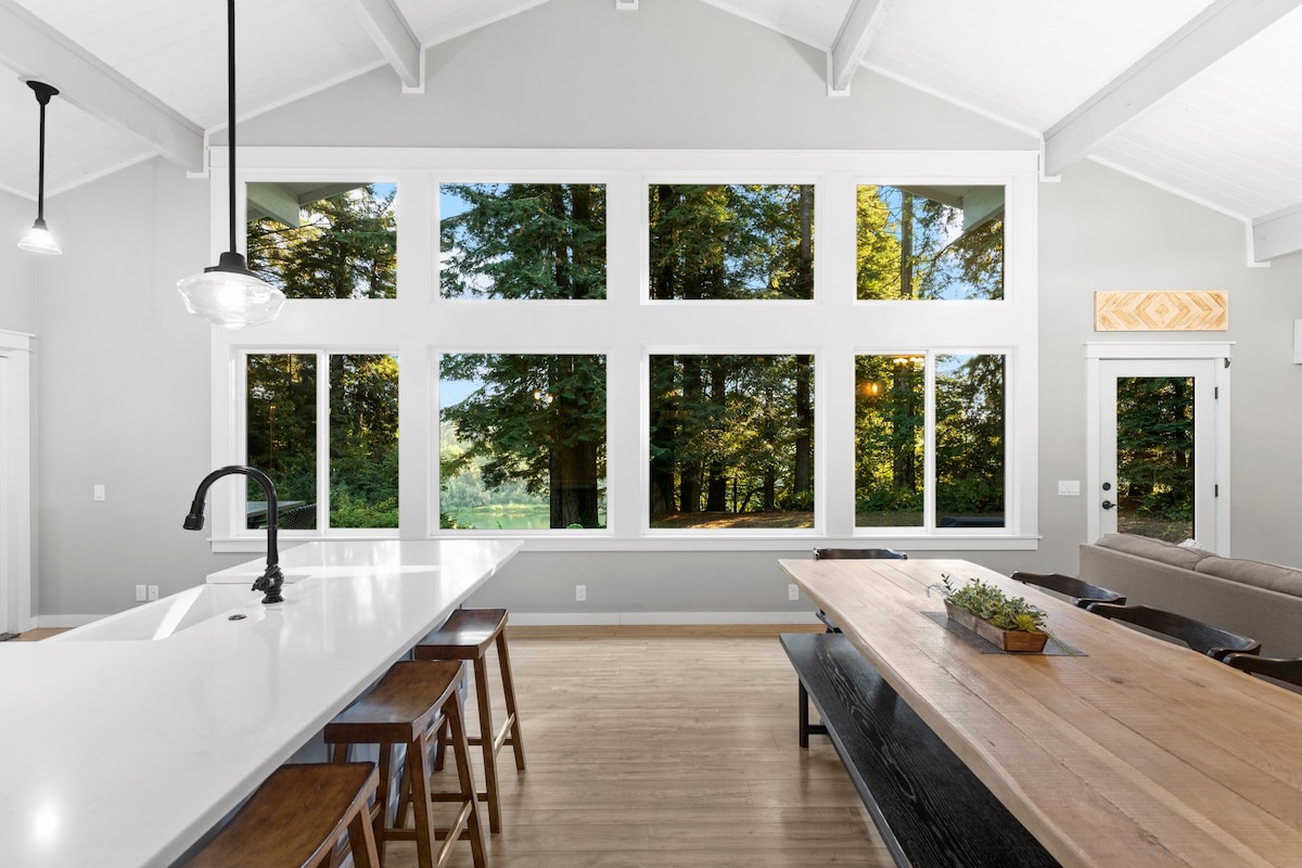 A modern kitchen and dining area features large windows offering views of the surrounding trees. The space is designed with a white countertop, dark wooden stools, and a dining table made of reclaimed wood. Natural light fills the room, creating an inviting atmosphere.