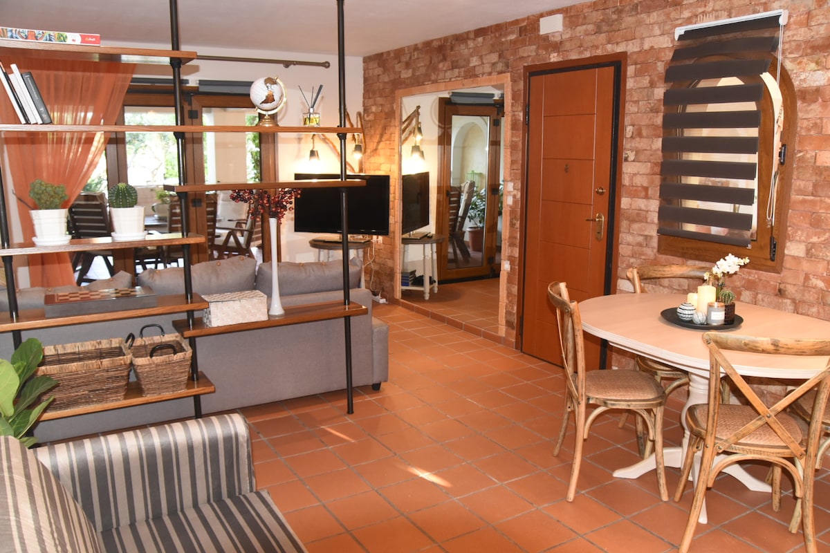 The interior space features warm brick walls and a cozy layout. A seating area includes a gray sofa and a striped armchair, while a round dining table is surrounded by wicker chairs. Large windows allow natural light to brighten the area, enhancing the inviting atmosphere.
