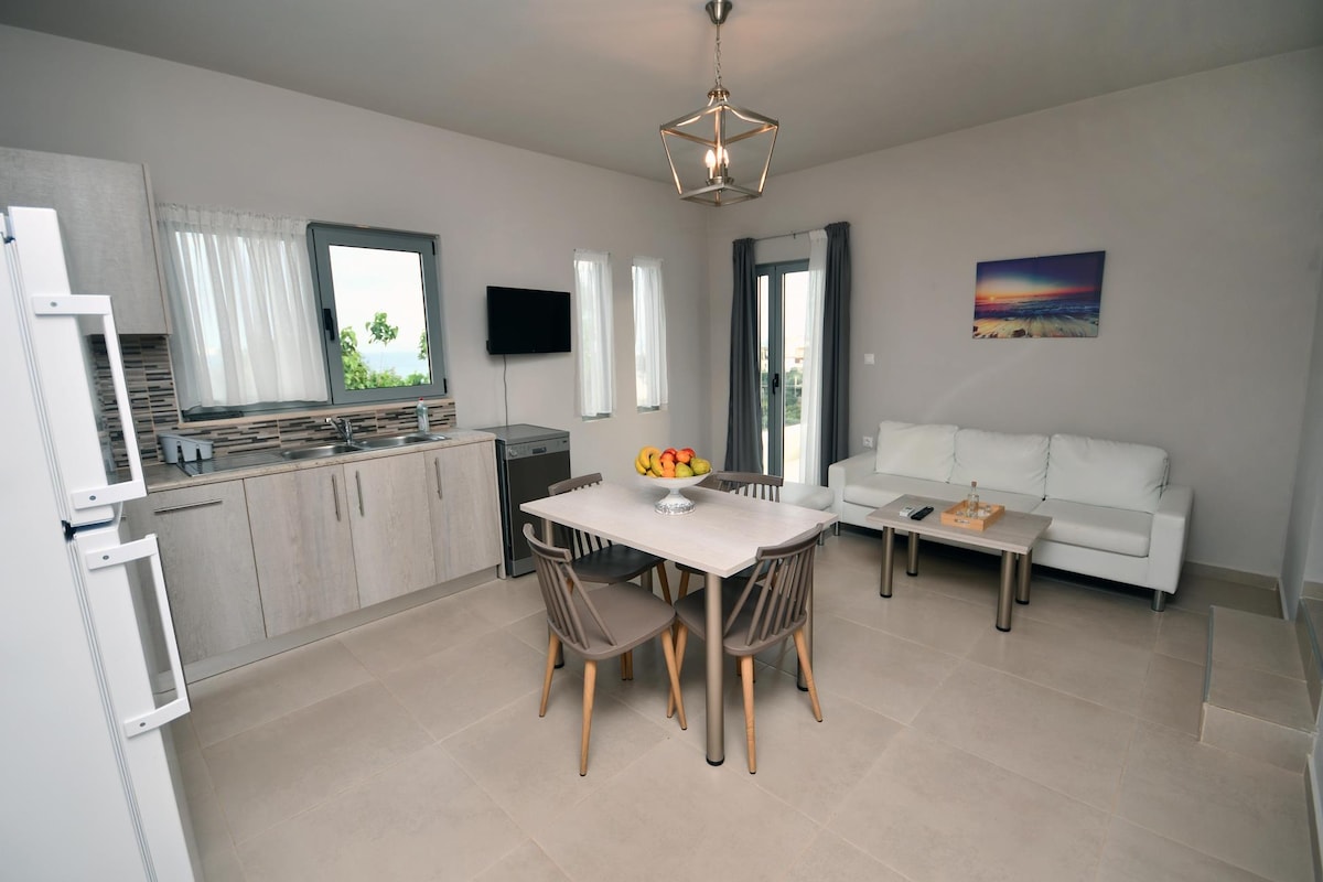 A bright and airy living space showcases a dining table surrounded by four light wooden chairs. A modern kitchen with grey cabinetry sits against one wall, while a white sofa invites relaxation. Large windows provide natural light and views of the outdoors.
