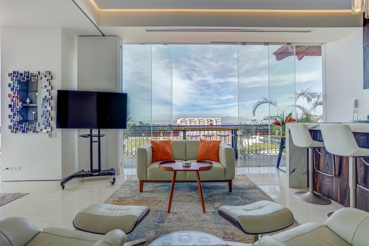 An open-concept living area is captured, featuring a modern design with a flat-screen TV and stylish seating arrangements. Floor-to-ceiling glass windows offer a view of the exterior, while the space is highlighted by soft LED lighting and elegant furnishings.