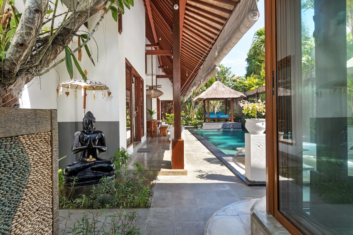 A serene pathway leads to a spacious private pool, surrounded by lush greenery. Traditional Balinese decorations, including a statue and gazebo, enhance the tranquil atmosphere. Large windows reflect natural light, connecting indoor and outdoor spaces seamlessly.