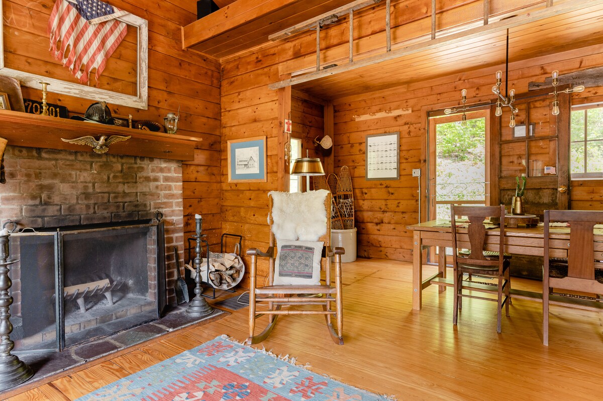 A rustic living space showcases warm wooden walls and a stone fireplace. Comfortable seating is provided by a rocking chair, while a dining area features a wooden table and chairs. Natural light filters through windows, highlighting the cozy atmosphere.