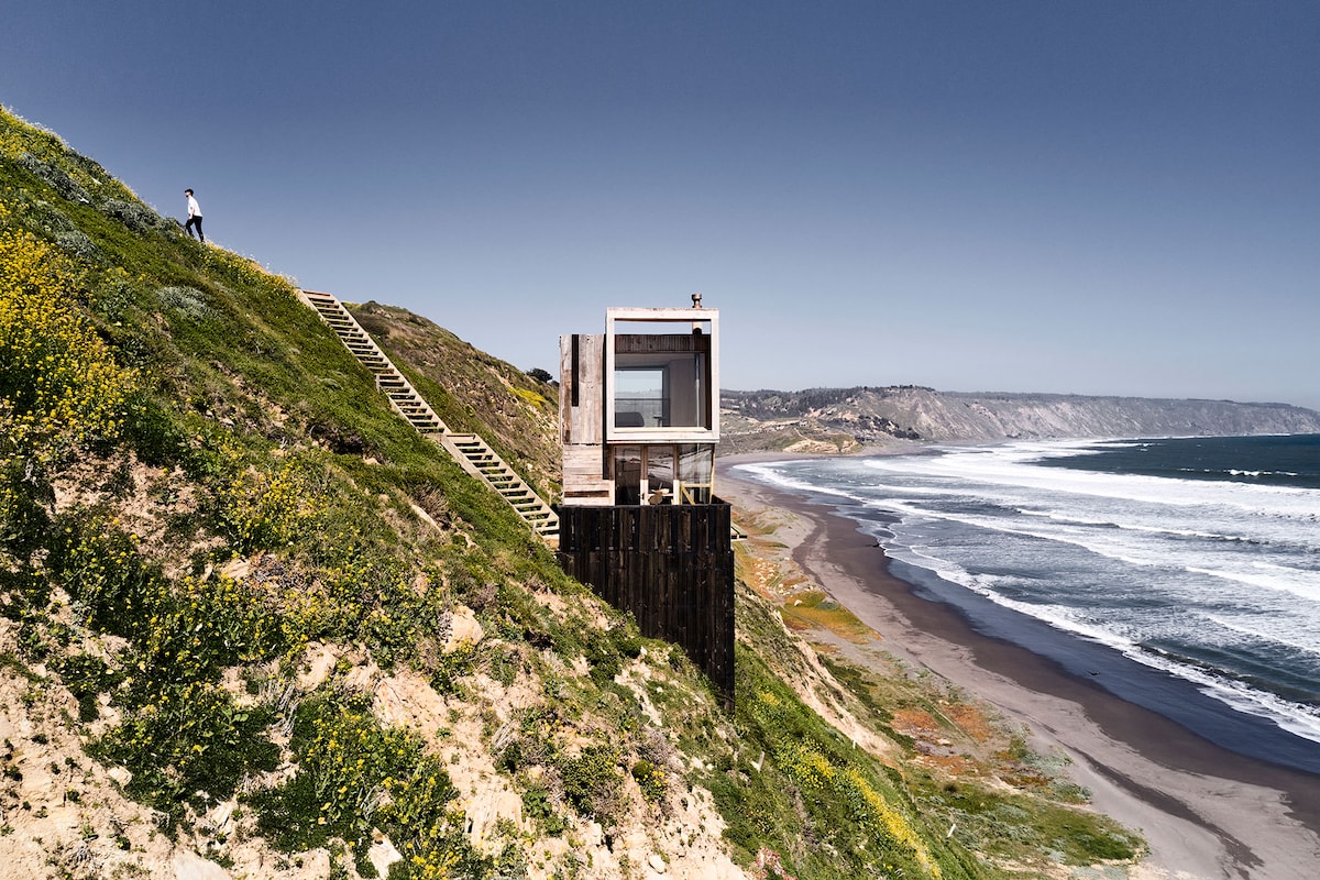 A cottage built on stilts overlooks the coastline, featuring large windows that offer an expansive view of the sea. Steps lead up the hillside to the entrance, while the surrounding area is adorned with vibrant wildflowers.
