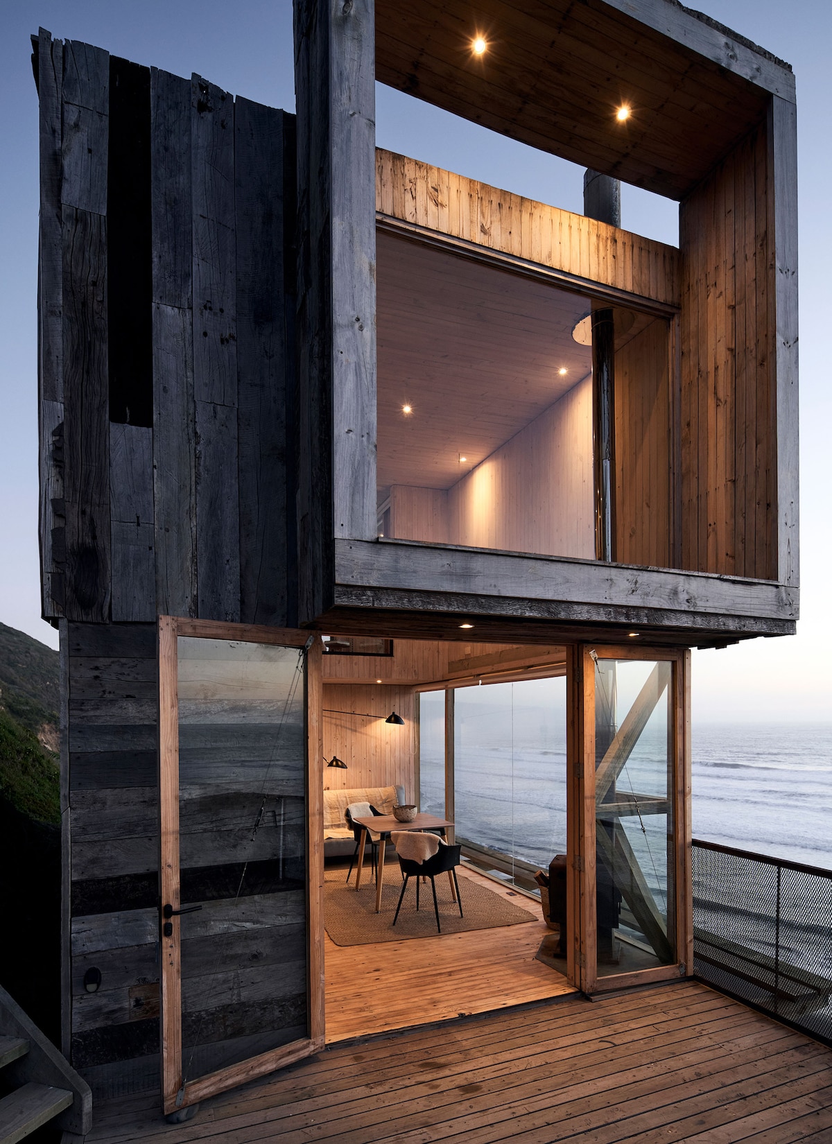 The exterior structure features a mix of rustic wood and modern design elements. Large glass doors open onto a deck that overlooks the sea. The warm wood interior is illuminated by soft lighting, creating a welcoming space with ocean views.