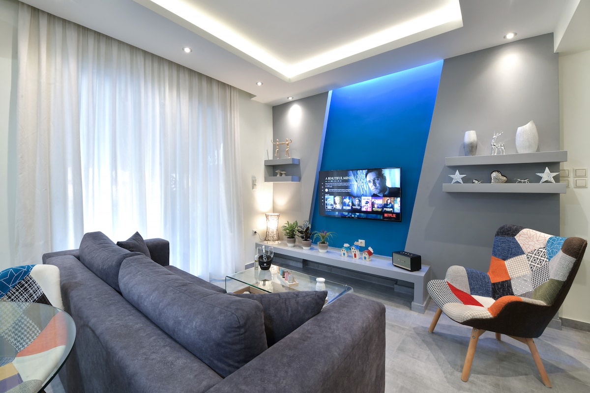 The living room features a large, gray sofa with colorful accent cushions. A 50-inch Android TV is mounted on a blue accent wall, complemented by decorative shelves with various items. Natural light filters through sheer curtains, creating a bright and welcoming space.