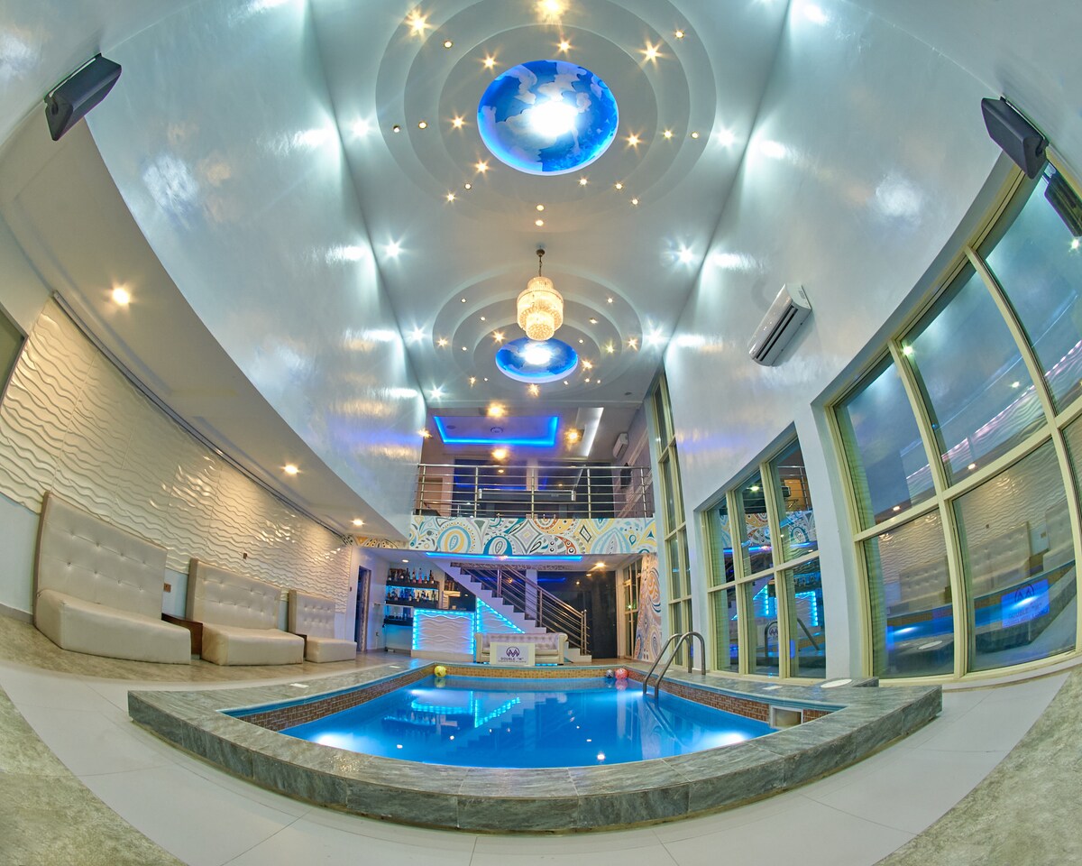 An indoor pool is highlighted in a modern lounge area, with soft seating arranged along the walls. Bright ceiling features include a chandelier and decorative lighting, creating an inviting ambiance. Large windows provide views of the exterior, enhancing the open and airy feel of the space.