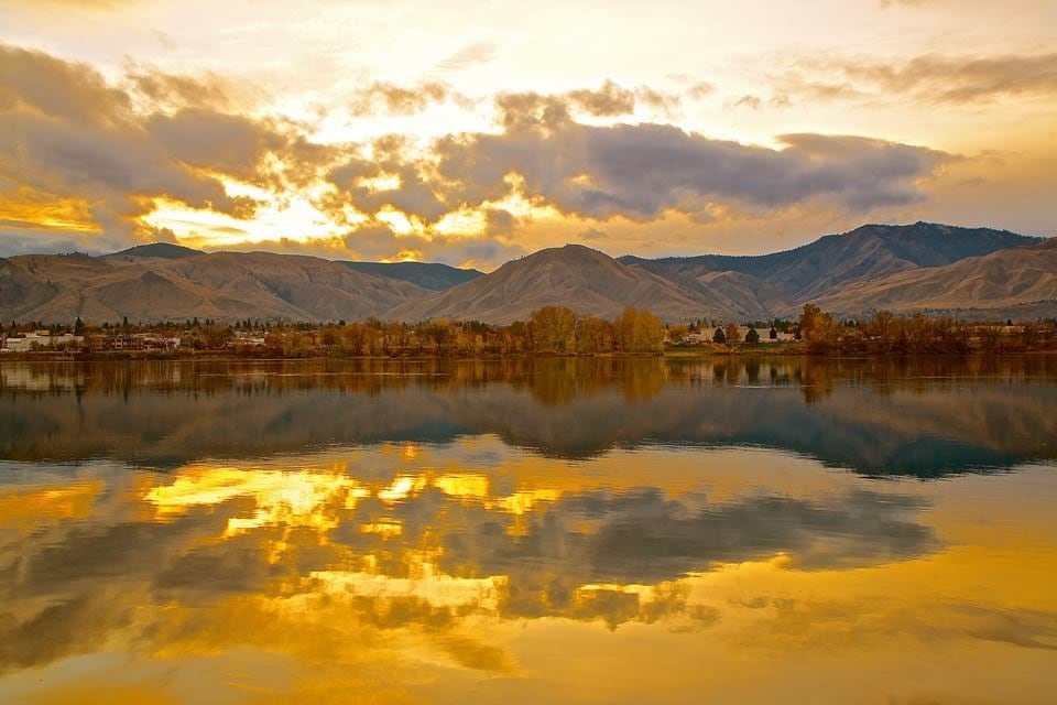A serene landscape captures the golden hues of sunset reflecting off the water's surface. The scene features rolling mountains in the background, framed by a soft palette of browns, golds, and subtle greens, creating a peaceful connection between sky and water.