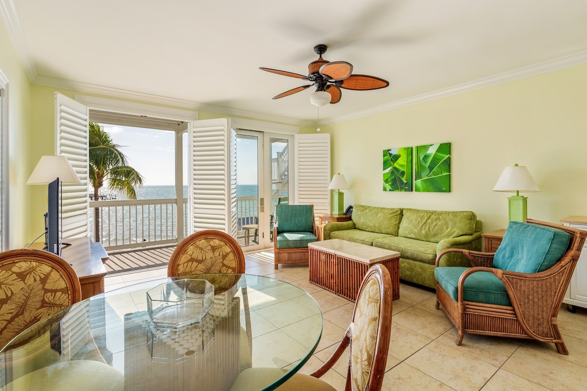 The living room features a spacious green sofa complemented by wicker armchairs. A glass dining table sits at the center, with light streaming through open shutters. The backdrop showcases ocean views, enhancing the connection to the balcony and natural surroundings.