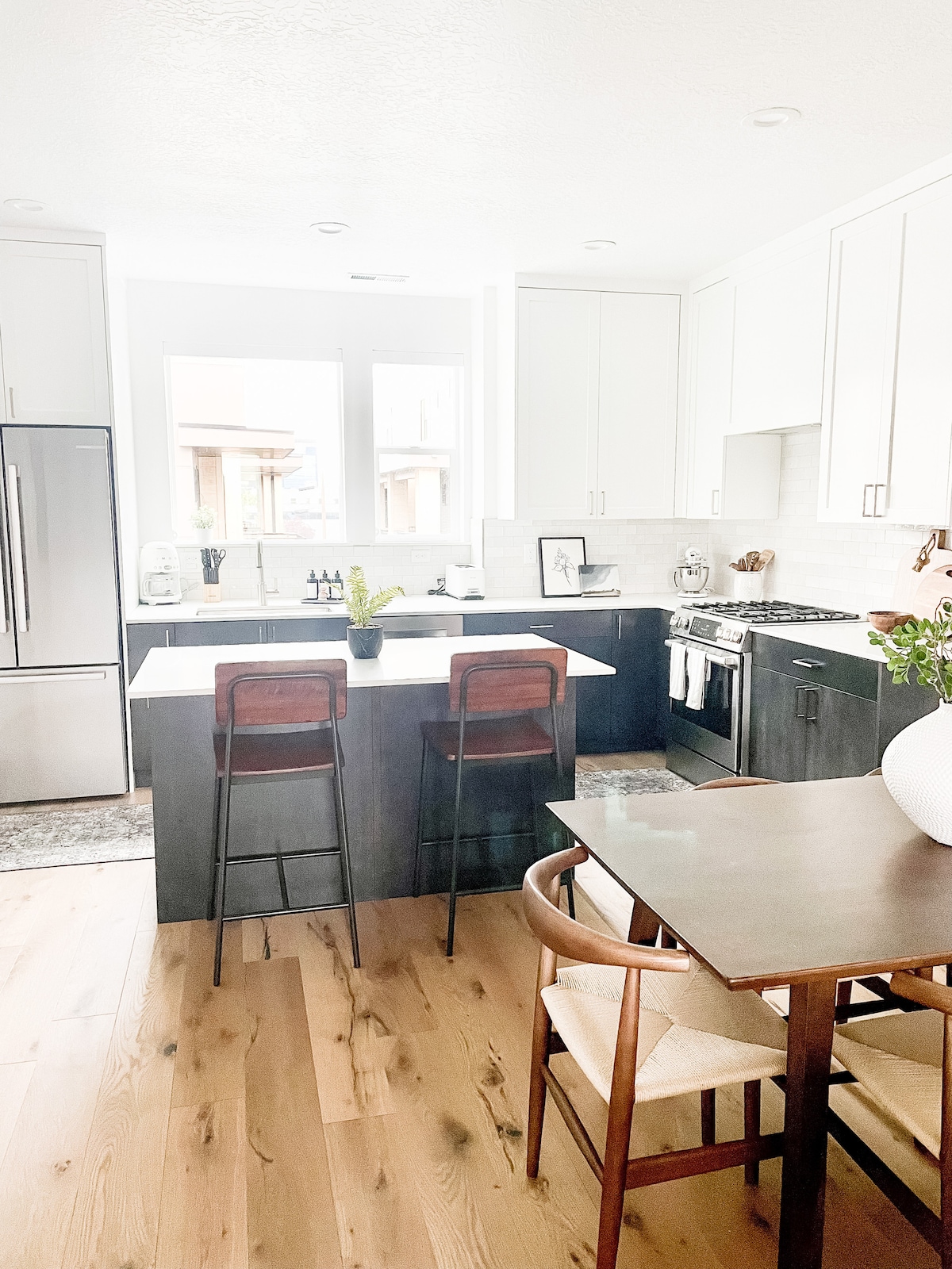 A modern kitchen features sleek cabinetry and stainless steel appliances. An island with two barstools stands at the center, complemented by a dining table with additional seating. Natural light enters through two windows, highlighting the open layout and warm wood flooring.