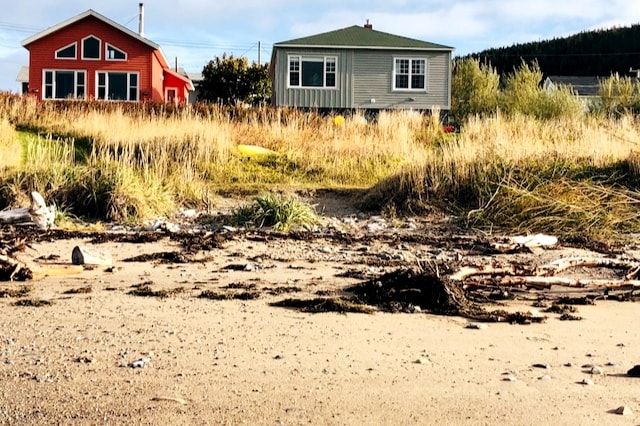 Two charming single-story homes are seen from the beach, framed by tall grass and sandy terrain. One house is painted in a warm red hue, while the other features a soft green exterior. The sandy beach is scattered with natural debris, adding to the coastal ambiance.