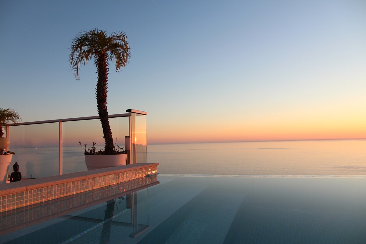 A serene salt-water pool is featured, blending into the horizon. A palm tree in a planter provides a tropical accent, while the sunset casts warm hues over the calm sea. The pool's edge meets the reflective water, creating a seamless connection with the sky.