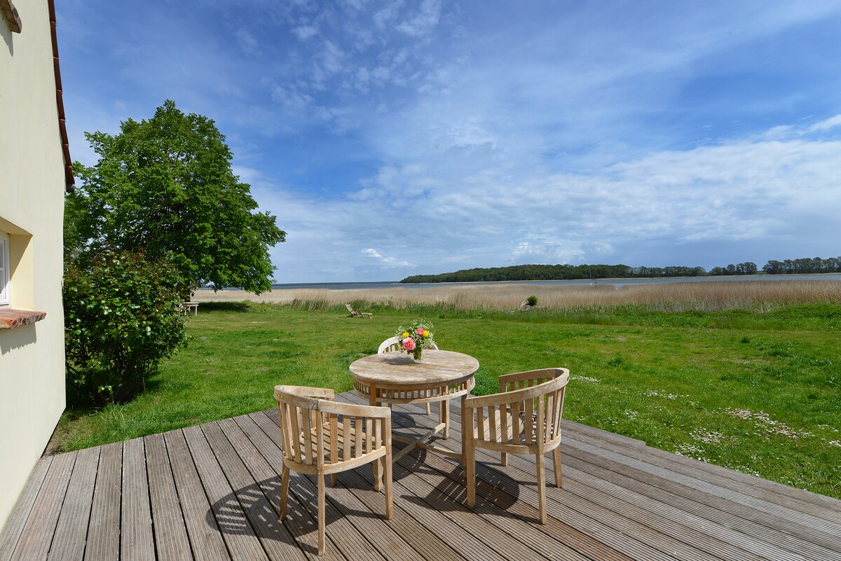 A wooden terrace hosts a round stone table surrounded by four wooden chairs. Lush green grass extends toward the water, with a view of a serene landscape under a partly cloudy sky. A tree provides shade nearby, enhancing the outdoor setting.