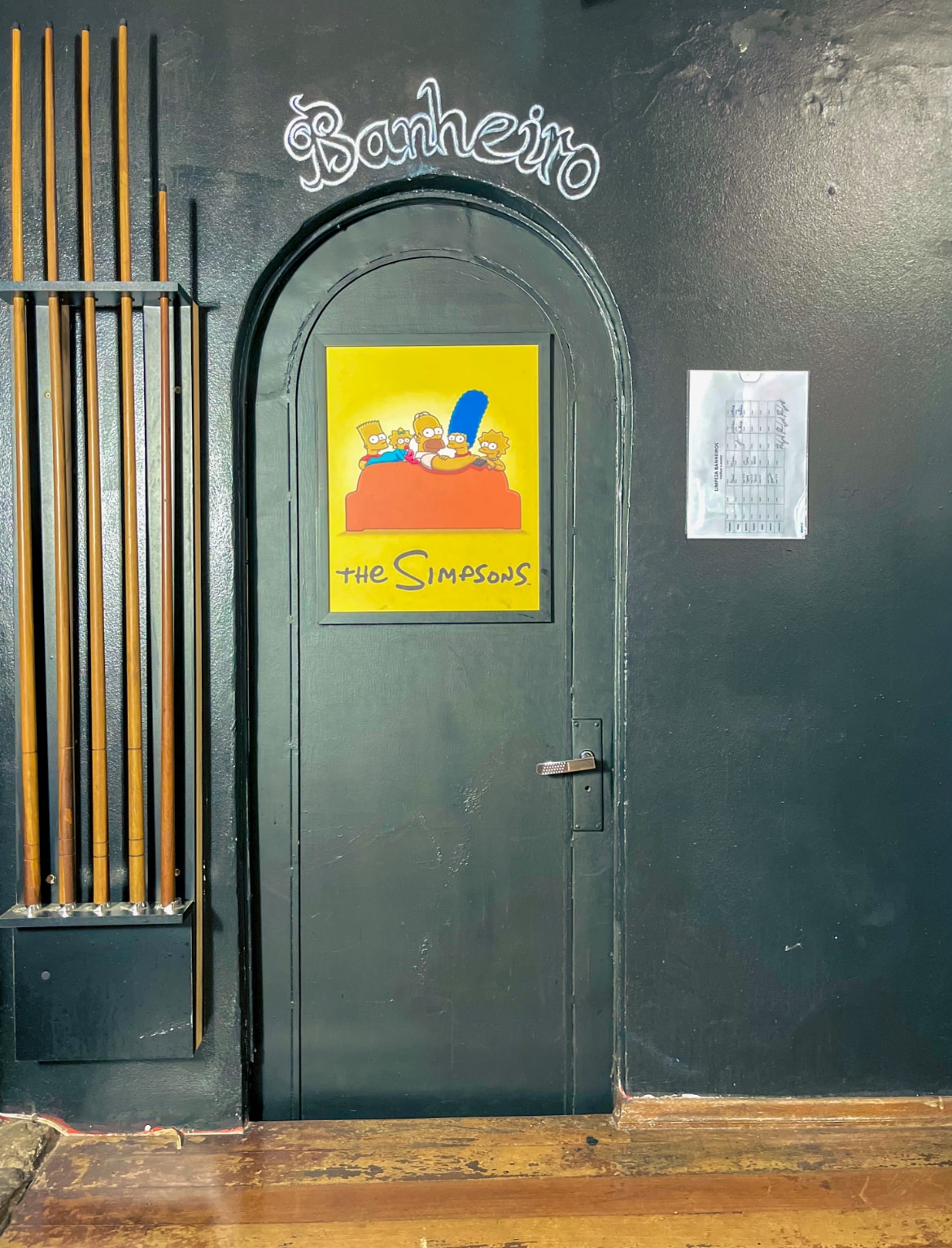 A black door with an arched top leads to the restroom, featuring a colorful poster of 'The Simpsons' displayed prominently. The door handle is metallic, and wooden slats can be seen to the left. A light-colored paper is taped to the wall nearby.