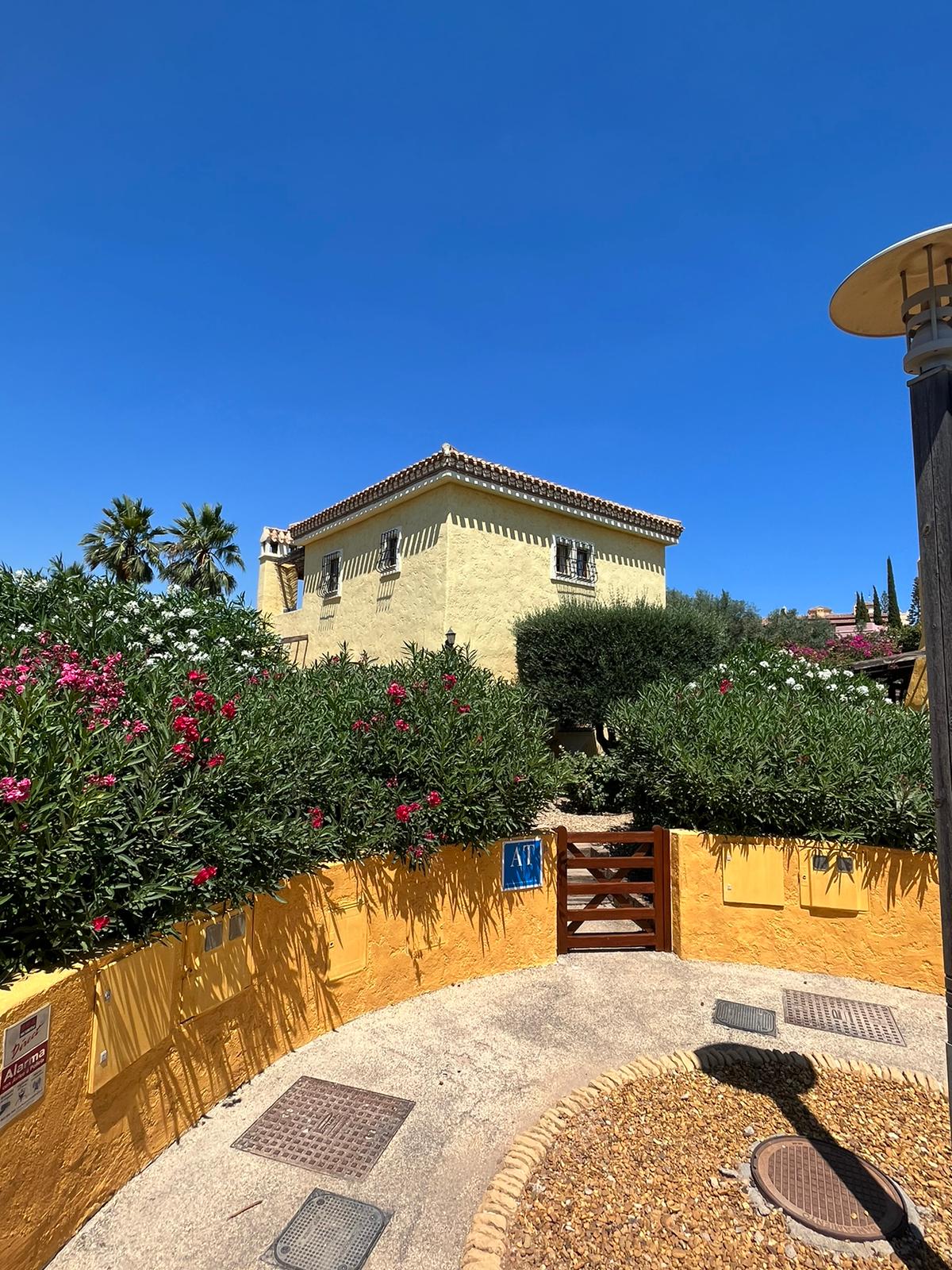 A charming villa is framed by lush greenery, showcasing vibrant flowering bushes in the foreground. The structure, featuring a traditional Spanish-style design, has a warm yellow exterior under a clear blue sky. An inviting pathway leads to a wooden gate, enhancing the outdoor atmosphere.