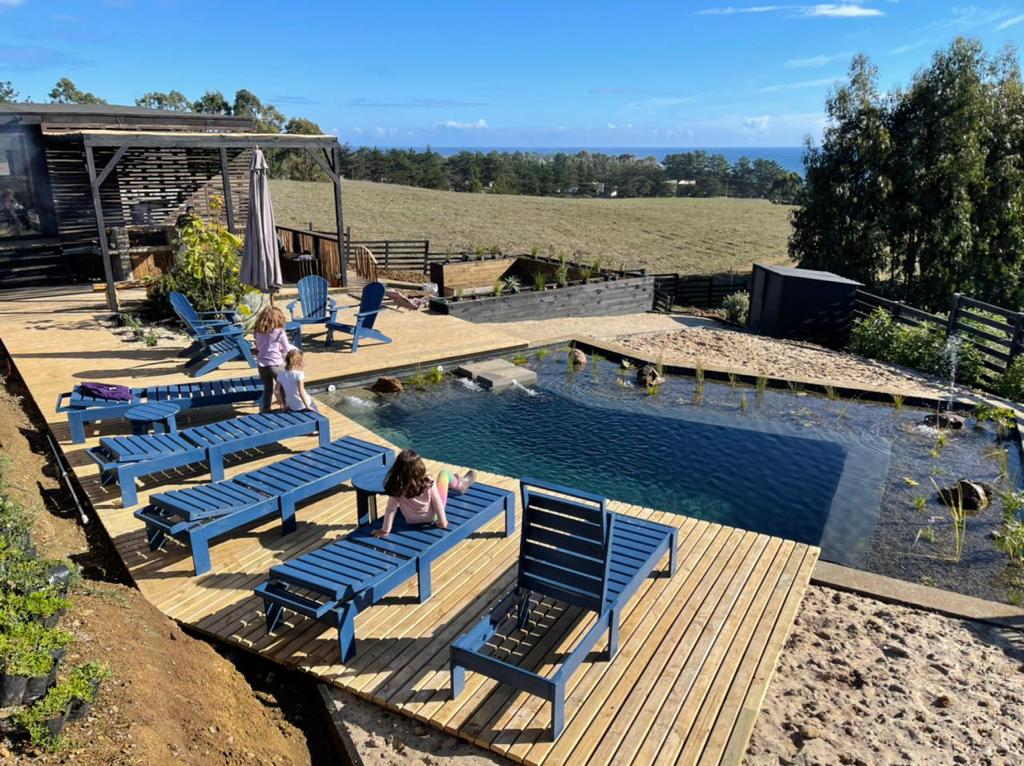 A natural swimming pool is surrounded by wooden decking, featuring several blue lounge chairs for relaxation. Lush landscaping and open fields with a distant view of the ocean contribute to a serene outdoor setting.