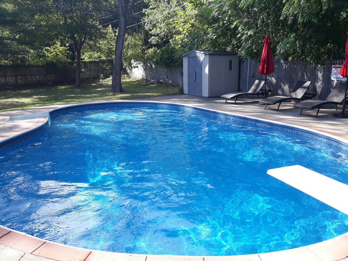 A heated in-ground swimming pool is surrounded by a spacious deck, featuring several lounge chairs oriented for relaxation. Lush greenery provides a natural backdrop, while red umbrellas offer shade nearby. A storage shed is visible in the corner, complementing the outdoor setting.