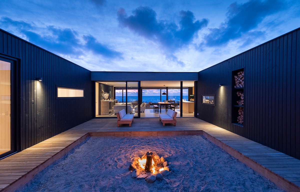 A modern outdoor area features a fire pit surrounded by sand, with two lounge chairs positioned nearby. The structure's sleek black exterior is complemented by large windows that offer views of the landscape. Dusk light casts a serene ambiance over the space.