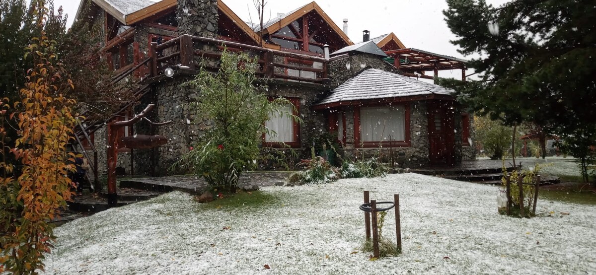 A rustic stone cabin is surrounded by a snowy landscape, with fresh snow covering the ground and the roof. The front terrace and garden are visible, featuring sparse greenery and subtle landscaping elements. Snowflakes can be seen falling gently in the scene.