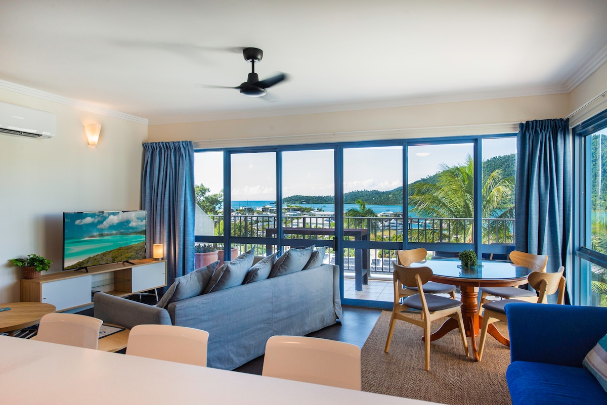 A light-filled living area features large windows offering panoramic views of the Whitsunday Seascape. A comfortable sofa sits beside a circular dining table, while a wall-mounted TV displays a coastal scene. Breezy curtains frame the windows, enhancing the inviting atmosphere.