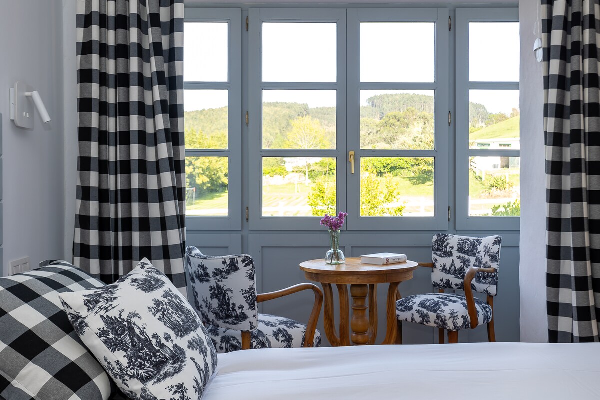 A relaxing room is presented, featuring large windows with a nature view and complemented by checkered curtains. A small wooden table is surrounded by two patterned chairs. Floral accents add a touch of color to the space, which is designed for comfort.