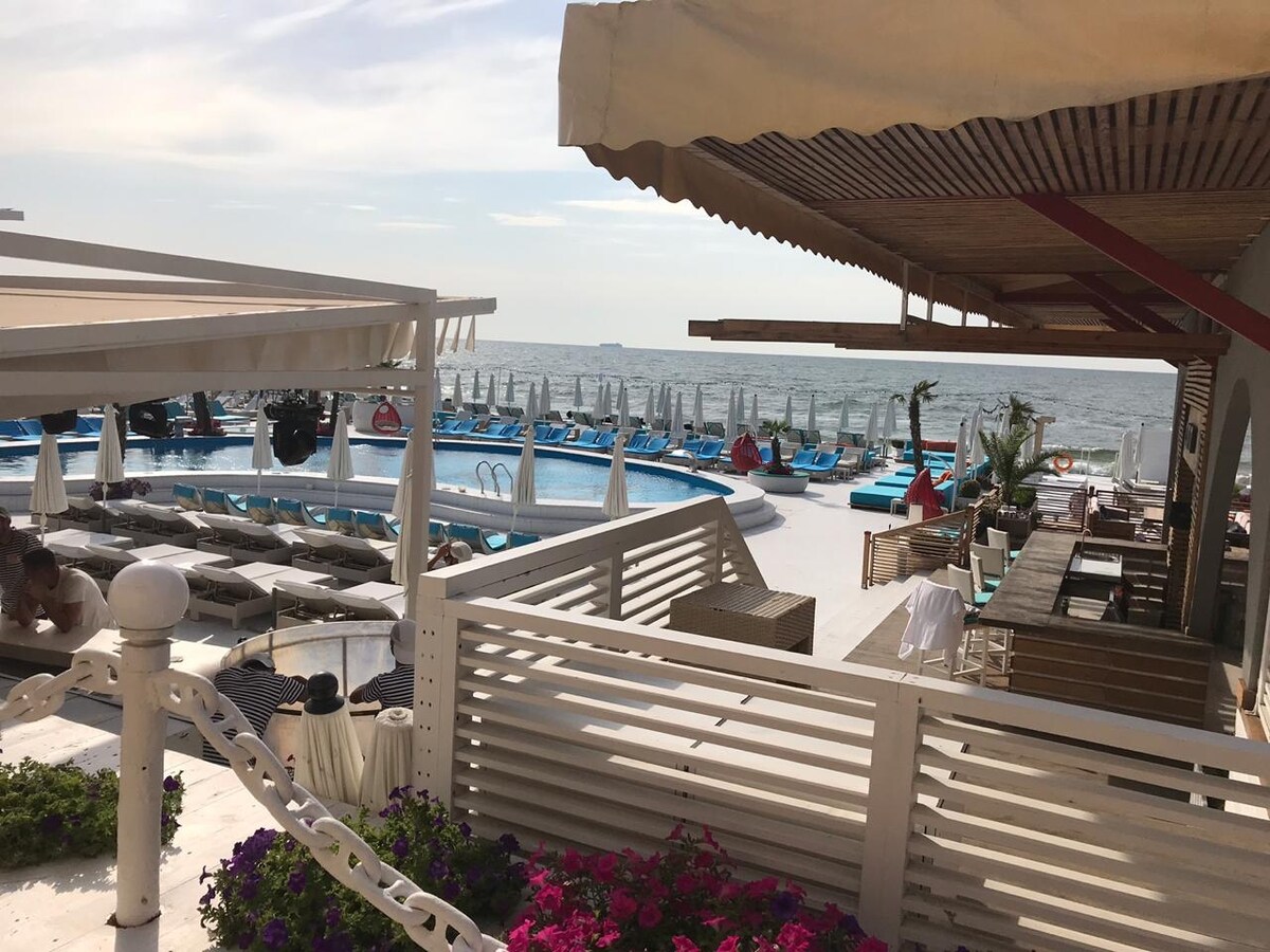 A seaside resort area is depicted, featuring a large pool surrounded by sun loungers. A shaded dining area with wooden tables and chairs overlooks the water, with umbrellas providing shade. Colorful flowers add vibrancy to the outdoor space, complementing the tranquil coastal scene.