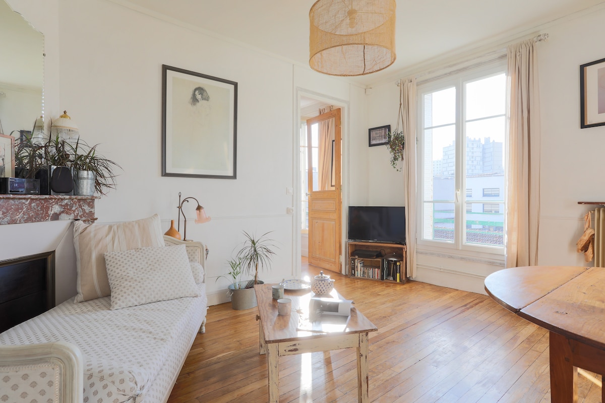 A bright living area features a mix of natural light from large windows, showcasing wooden floors and cozy furnishings. A light-colored chaise lounge sits beside a low table, while a television is positioned on a shelf. Decorative plants enhance the homey ambiance.