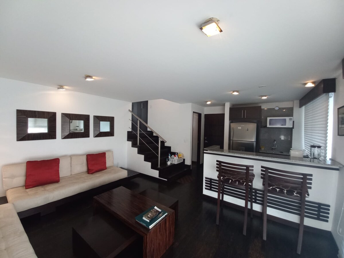 A modern living area features a light-colored sectional sofa adorned with red pillows, complemented by a dark wooden coffee table. A bar counter with two chairs separates the living space from a well-equipped kitchen. The staircase leads to upper levels, enhancing the open design.