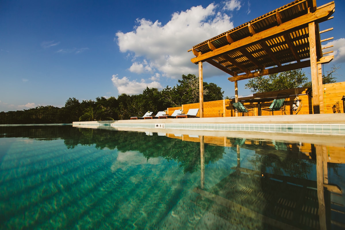 A serene infinity pool reflects the clear blue sky and surrounding lush greenery. A shaded wooden pergola provides relaxation space with several lounge chairs positioned nearby, inviting guests to unwind in nature's tranquility.