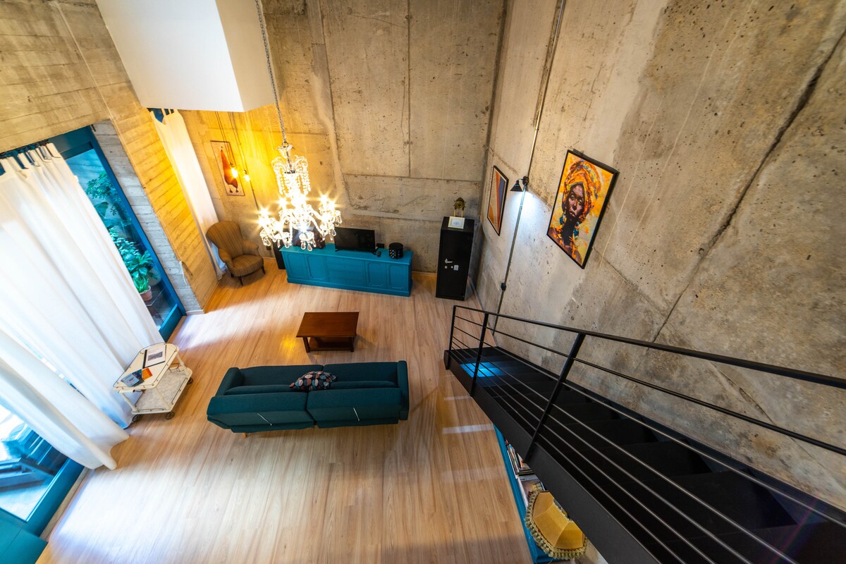 An open and airy loft space is highlighted by tall concrete walls and large windows, allowing natural light to flood the room. A spacious green sofa sits opposite a wooden coffee table, while a modern chandelier hangs from the ceiling, adding character to the design.