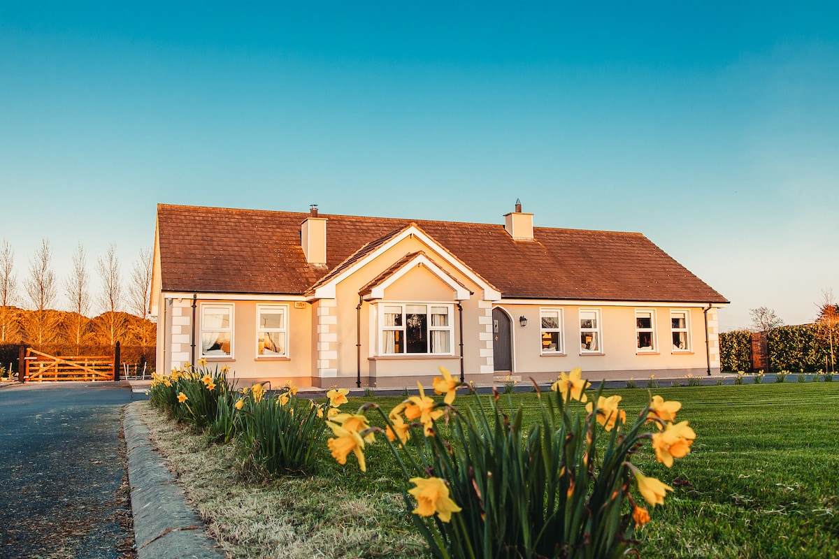 A charming bungalow is set against a clear sky, featuring a warm exterior in soft tones. Bright yellow daffodils bloom in the foreground, enhancing the welcoming entrance. The spacious lawn surrounds the home, with neatly trimmed hedges providing natural borders.
