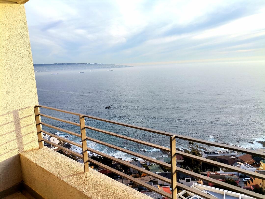A panoramic view of the ocean is showcased from the terrace, where the sea meets the horizon. The image captures the gentle waves and distant ships, with the sky reflecting soft hues of blue and gray. A clean railing frames the scenic overlook.
