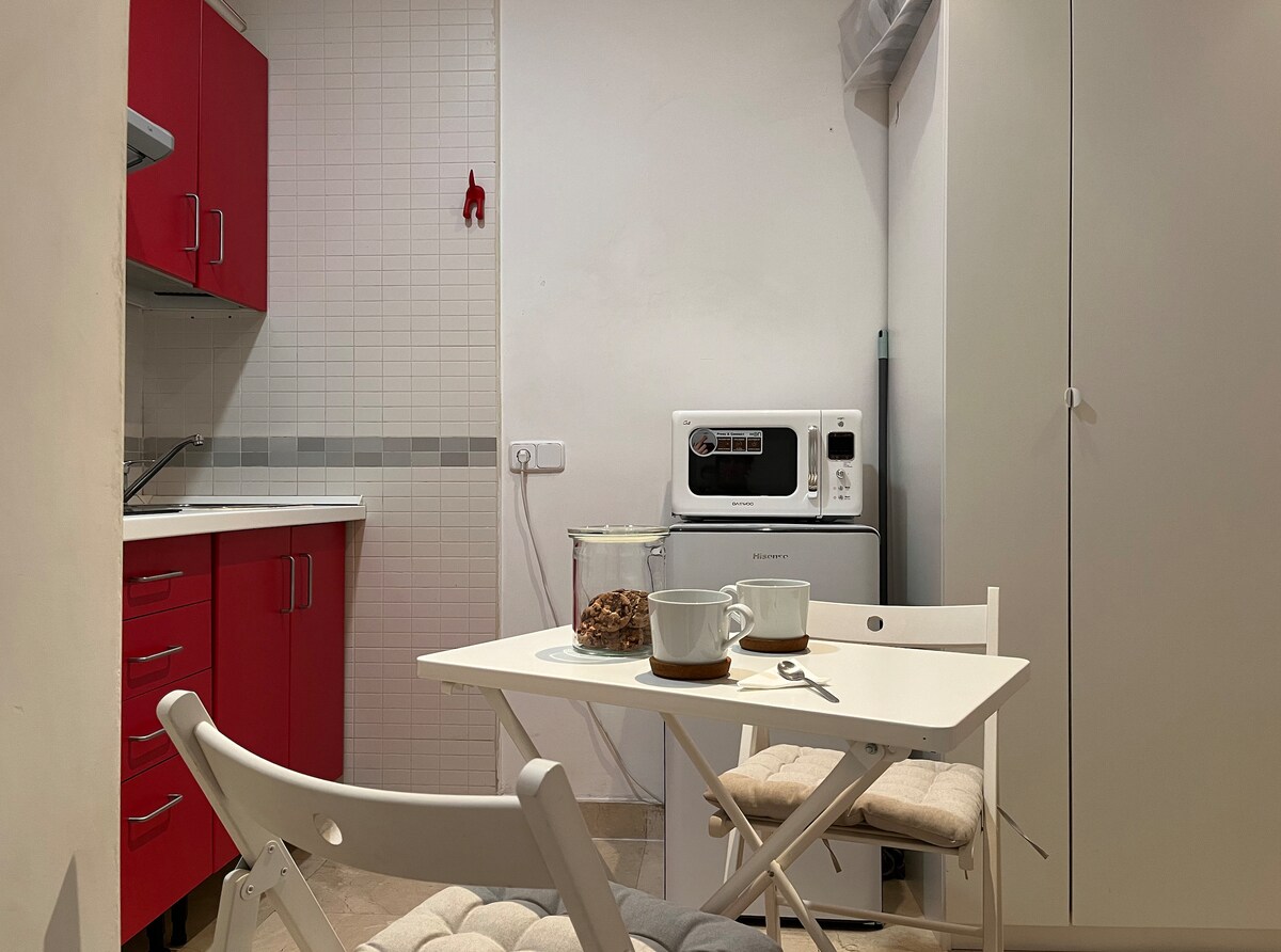 A compact kitchen area features red cabinetry and essential appliances, including a microwave and refrigerator. A small, white dining table is set for two, complemented by light-colored padded chairs. The walls are finished in a neutral tone, creating a functional yet welcoming space.