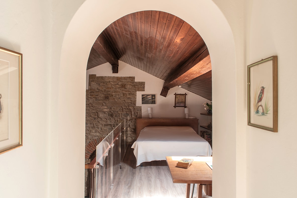 The image showcases a loft area framed by an archway, featuring a spacious bed and wooden ceiling beams. A wooden table is visible nearby, and stone walls add texture to the cozy atmosphere. Soft lighting enhances the intimate setting.